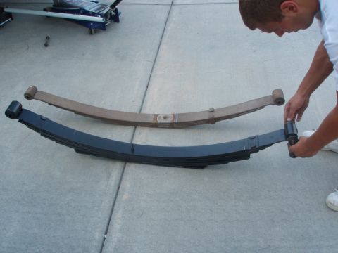 1987 Ford Bronco - A person holds a black leaf spring assembly next to a rusty leaf spring assembly, both laid out on a concrete driveway.