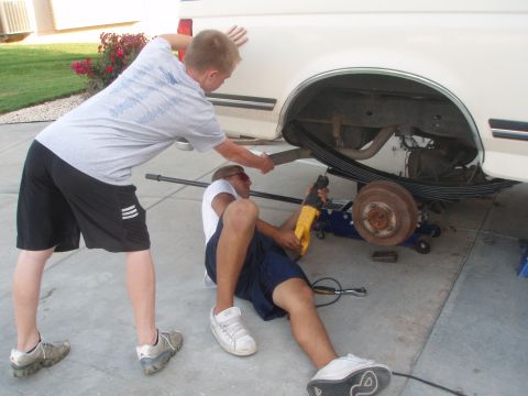 1987 Ford Bronco - Two men work on the rear suspension of a white pickup truck, with one using a reciprocating saw near the leaf springs and drum brake.