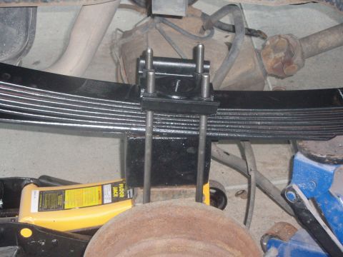 1987 Ford Bronco - A close-up view of the underside of a vehicle, focusing on a leaf spring assembly with a U-bolt clamp. A yellow floor jack is visible suppo…