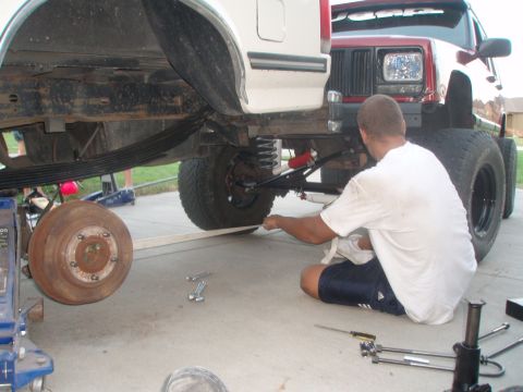 1987 Ford Bronco - A man in a white t-shirt and blue shorts works on the rear suspension of a lifted white pickup truck, which is parked next to a red Jeep Ch…