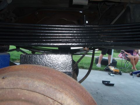 1987 Ford Bronco - The underside of a vehicle shows a black leaf spring assembly, a rusty brake rotor, and a black painted differential housing. A blue jack s…