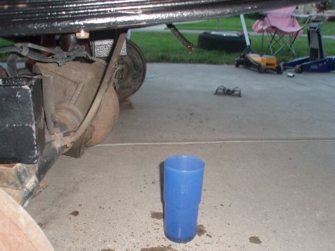 1987 Ford Bronco - The underside of a vehicle's rear axle assembly is shown, with a blue plastic cup placed on the concrete below. Various automotive tools an…