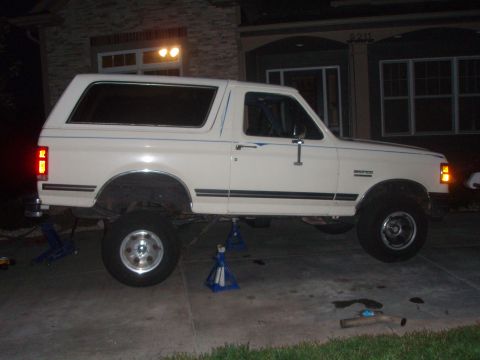1987 Ford Bronco - A white 1987 Ford Bronco is parked on a concrete driveway in front of a house, supported by jack stands. The vehicle appears to be undergoi…