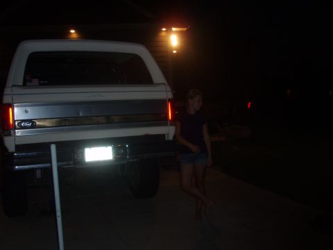 1987 Ford Bronco - The rear of a white Ford Bronco is visible at night, parked on a paved surface. A woman stands to the right of the vehicle.