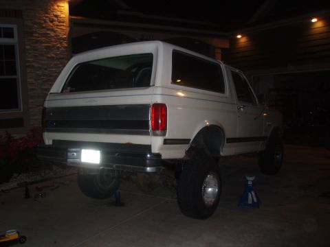 1987 Ford Bronco - The rear passenger side of a white Ford Bronco is shown lifted on jack stands at night. The vehicle has large aftermarket tires and is park…