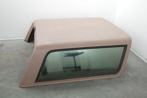1996 Ford Bronco - Photograph of a tan fiberglass truck cap with a glass window sitting on the floor, not a document.