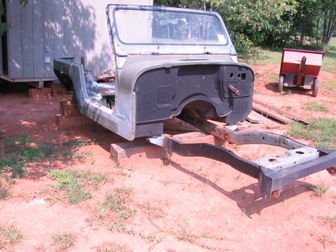 1980 Jeep CJ7 - The body and frame of a Jeep CJ7 are disassembled and supported by wooden blocks outdoors. The body is partially painted green and black, w…