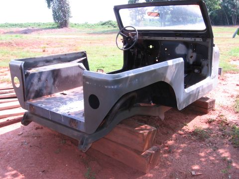 1980 Jeep CJ7 - The body of an olive green Jeep CJ7 is shown disassembled and resting on wooden blocks outdoors. The windshield frame, steering wheel, and…