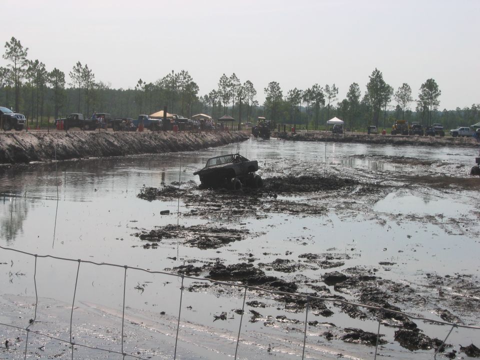 Dark 4x4 stuck in a mud-bog water pit with spectators and tents on the far bank