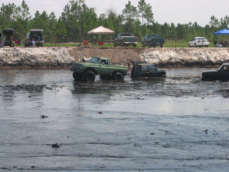 Off-road trucks and SUVs driving through a muddy water obstacle.