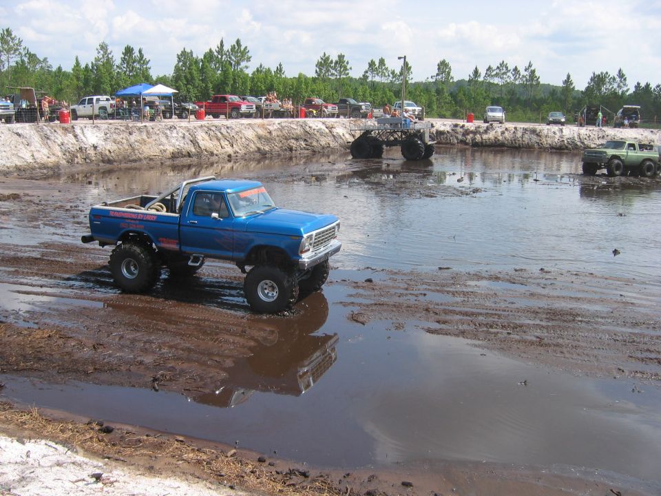 Lifted blue 1970s Ford pickup on mud tires in a red clay mud pit at an off-road event