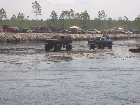 1992 Ford Bronco - Off-road vehicles navigate a muddy track during an event, with trees and tents visible in the background.