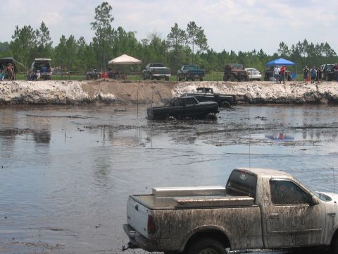 1992 Ford Bronco - Multiple trucks are stuck in a muddy, water-filled area while spectators watch from the bank.