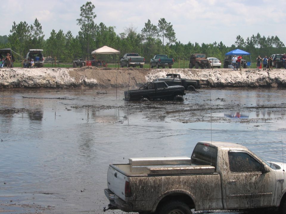Muddy white pickup truck in foreground, other trucks stuck in mud and water, spectators on a dirt bank