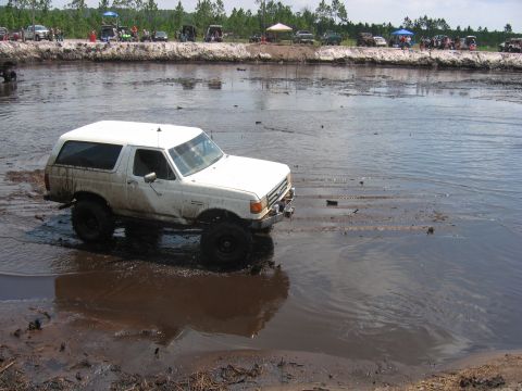 1992 Ford Bronco - A white Ford Bronco navigates a muddy off-road course, with spectators gathered on the embankment.