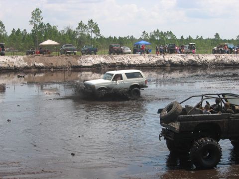 1992 Ford Bronco - A white Ford Bronco navigates a muddy track, with another modified off-road vehicle visible in the foreground.