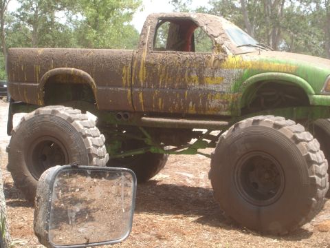 1989 Ford Bronco - A heavily muddied pickup truck with massive tires sits off-road, its green paint partially visible.