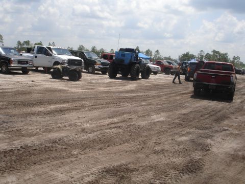 1989 Ford Bronco - A collection of trucks and a large blue tractor are parked in a muddy field under a cloudy sky.