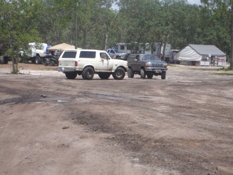 1989 Ford Bronco - Two SUVs are parked on a dirt lot, with trees and trailers in the background.
