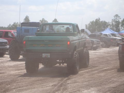 1989 Ford Bronco - A lifted Ford pickup truck is parked in a dusty outdoor area with other vehicles and tents visible.