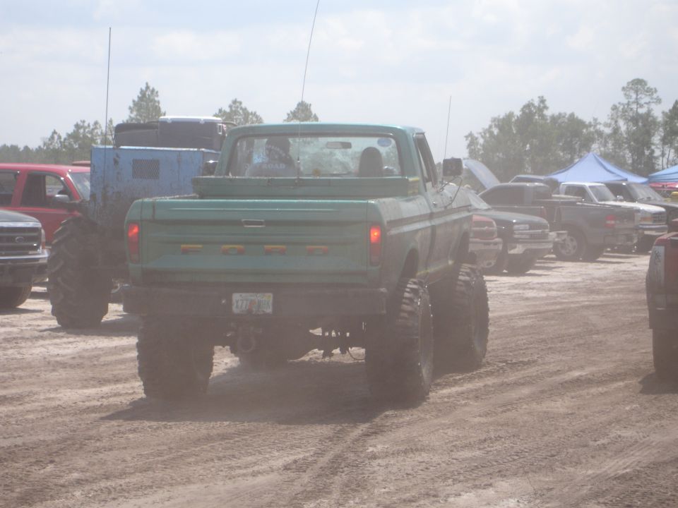 Green Ford pickup truck with large tires, seen from rear. Other trucks and tents in background