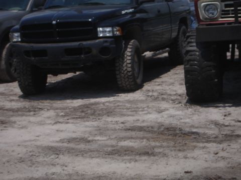 1989 Ford Bronco - Two lifted trucks, a black pickup and a red SUV, are parked on sandy ground.