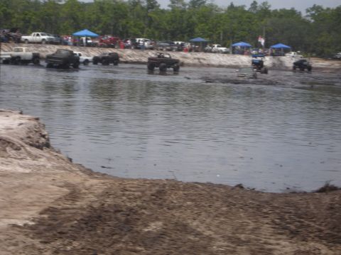 1989 Ford Bronco - Off-road trucks participate in a mud bog event with spectators watching from the shore.