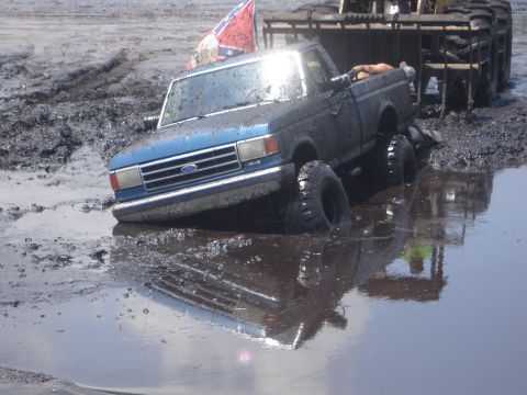 1989 Ford Bronco - A blue Ford pickup truck is stuck in a mud pit, with a large loader vehicle behind it.