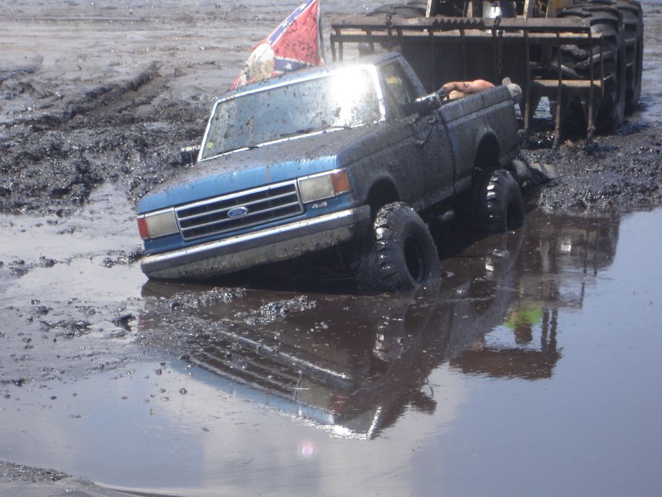 Ford pickup stuck deep in mud, with a loader behind it. Confederate flag on antenna