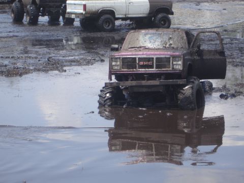 1989 Ford Bronco - A muddy GMC pickup truck sits in water with its door open, while other trucks are visible in the background.