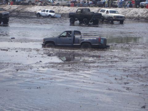 1989 Ford Bronco - A dark gray pickup truck is stuck in deep mud, with other vehicles and spectators in the background at an off-road event.