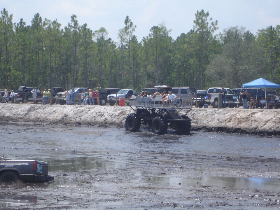 Monster truck with people riding in the back driving through a mud pit with other trucks and people on the bank