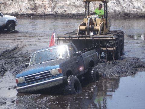 1989 Ford Bronco - A blue Ford pickup truck is being towed from a muddy area by a front-end loader, with another white truck in the background.