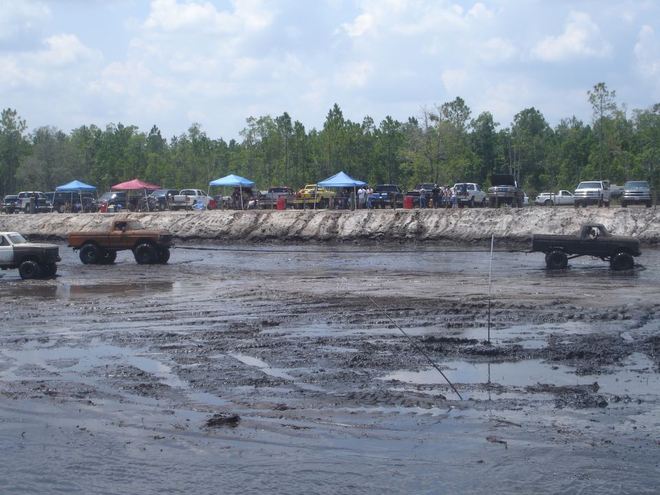 Trucks driving through a mud pit with spectators and tents in the background