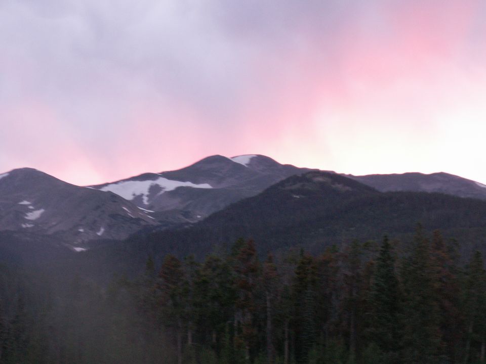 Mountains with snow patches under a pink and purple sky at dusk. Trees in the foreground
