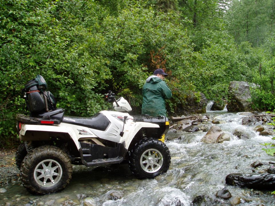 White all-terrain vehicle with luggage on the back, parked in a shallow stream with trees and rocks in the background
