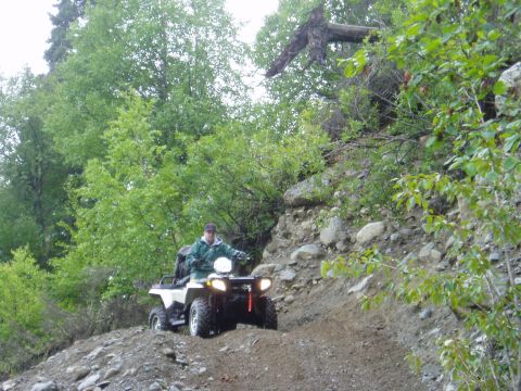 2000 Ford E350 - A person rides an all-terrain vehicle up a steep, rocky incline surrounded by trees.
