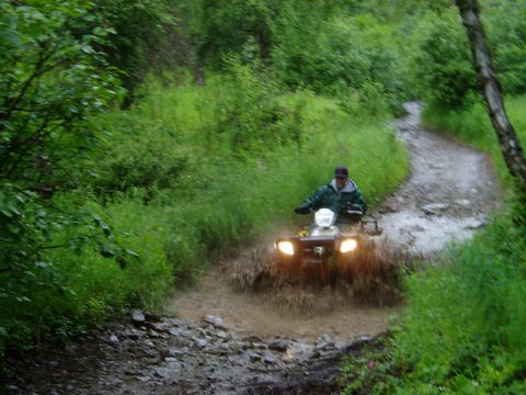 2000 Ford E350 - A person rides an ATV through a muddy trail surrounded by lush green foliage.
