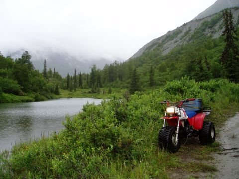 2000 Ford E350 - Three-wheeled ATV parked on a dirt path beside a lake with a wooded mountain in the background.