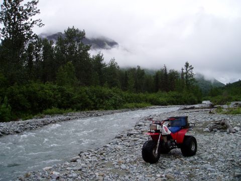 2000 Ford E350 - A red and white three-wheeled ATV is parked on a rocky riverbed next to a flowing river. The ATV has a blue storage box mounted on the rear…