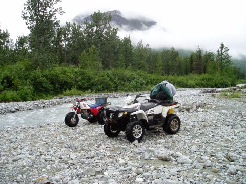 2000 Ford E350 - Two ATVs are parked on a rocky riverbed with a forest and misty mountains in the background. One ATV is red and white, and the other is whi…