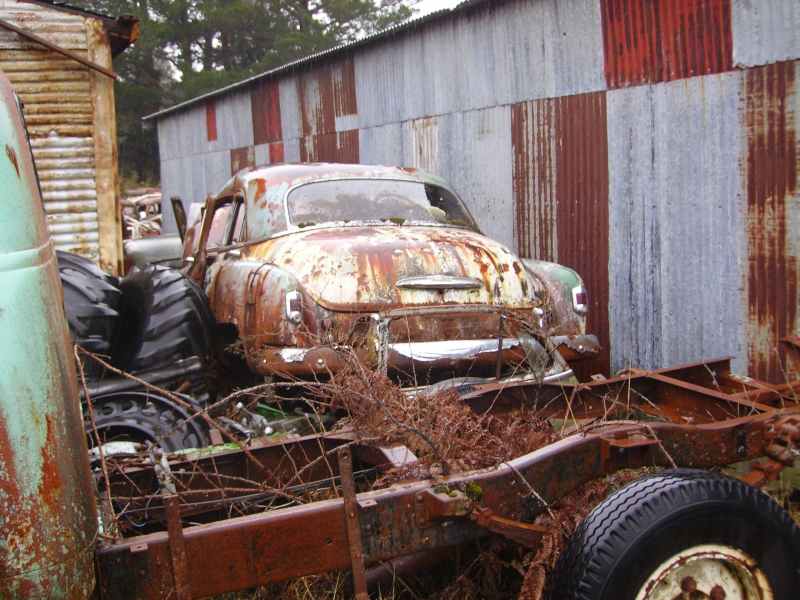 Rusted 1940s-50s fastback coupe wreck in a scrapyard beside a bare truck chassis