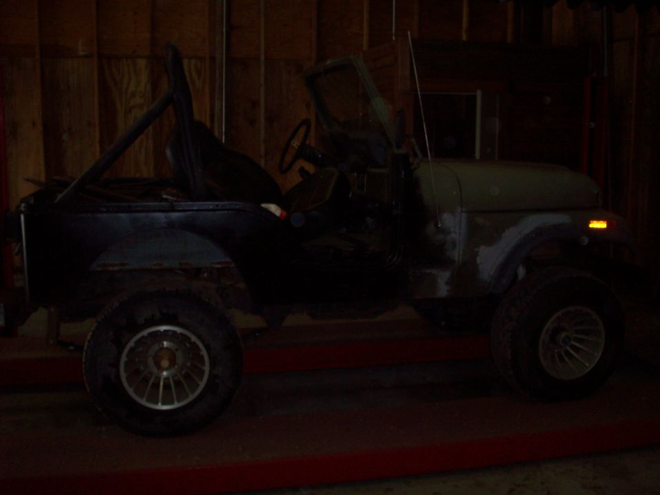 Side view of a dark Jeep CJ5 with a roll cage and green hood indoors.