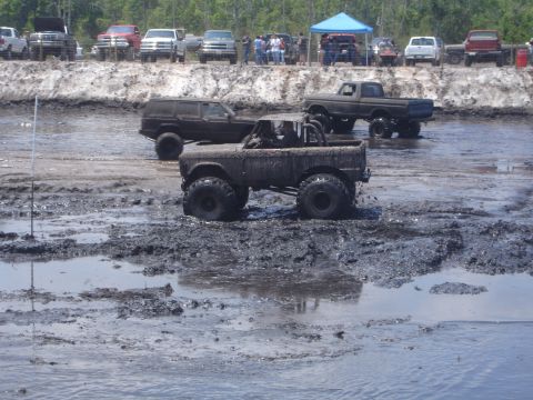 1989 Ford Bronco - A heavily mud-covered Ford Bronco is stuck in a mud pit, with a lifted black Jeep Cherokee and a dark-colored pickup truck in the backgroun…