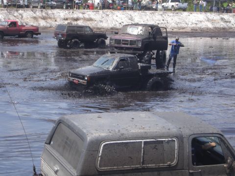 1989 Ford Bronco - A lifted, dark-colored pickup truck is driving through a muddy track, splashing water. A person is standing on the back of another lifted t…