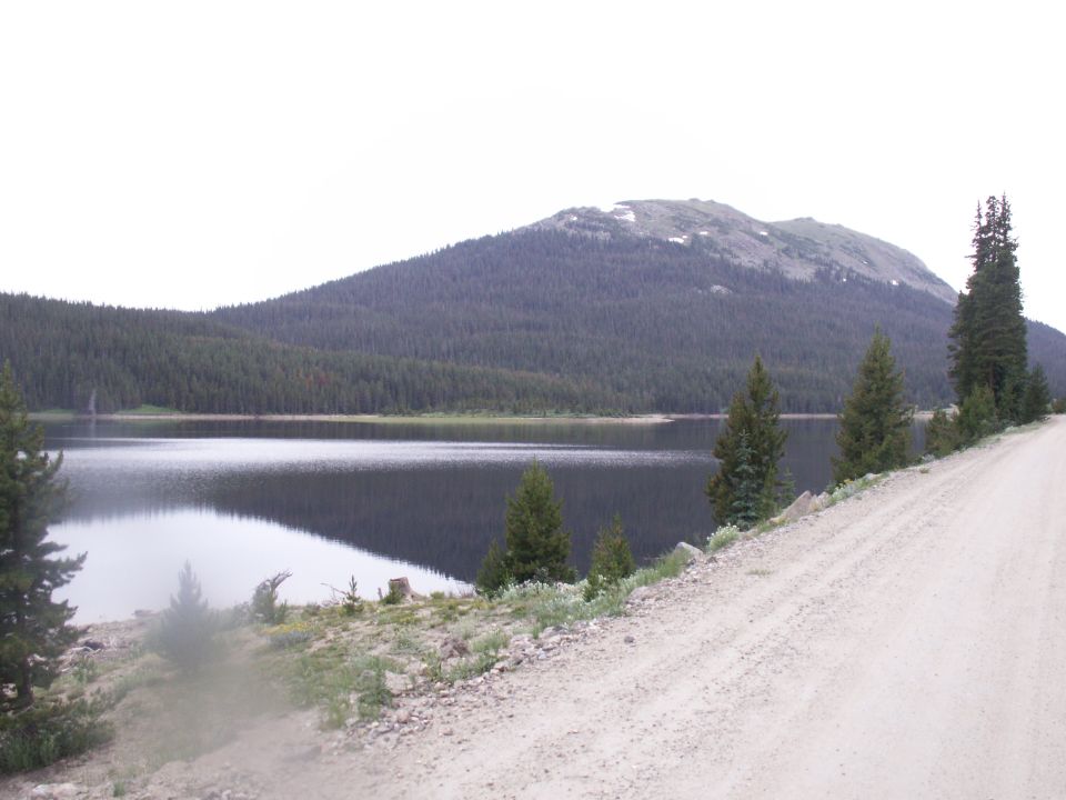 Dirt road next to a lake with a forest and mountain in the background.