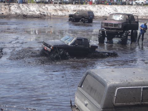1989 Ford Bronco - A black pickup truck is navigating through deep mud and water, creating a large splash. In the background, a lifted GMC pickup truck with l…