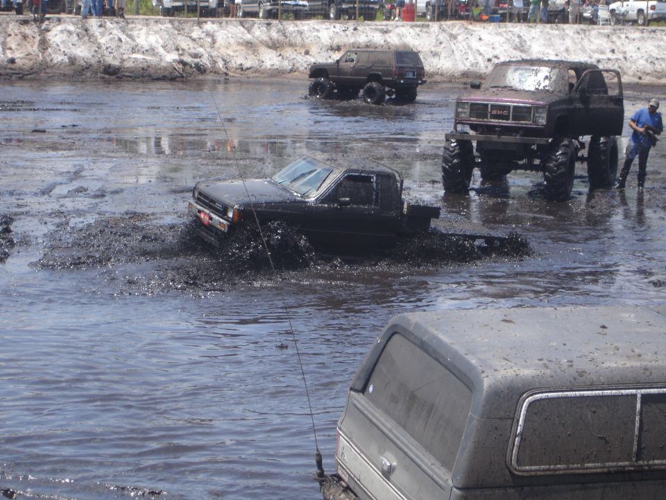 Black pickup truck driving through mud and water with a lifted GMC truck in the background.