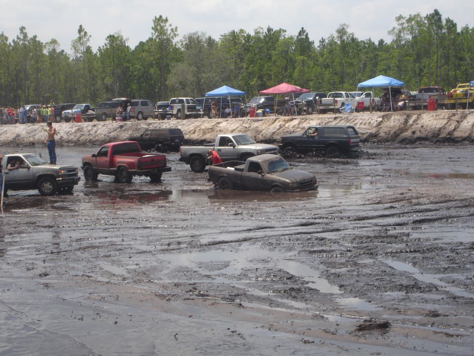 Dark SUV driving through mud pit with other vehicles in background.