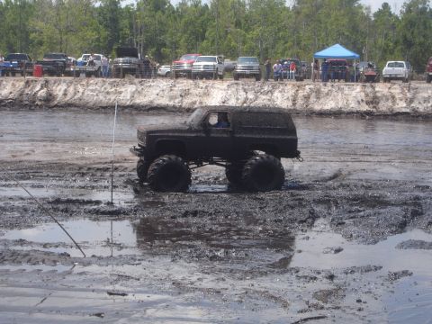 1989 Ford Bronco - A dark-colored, lifted SUV with oversized tires is driving through a muddy track. Several other vehicles and spectators are visible on a ra…